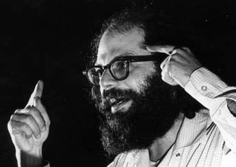 A photo of Ginsberg speaking somewhere, his hands are out in front of him with his fingers pointing sideways diagonally towards the audience as if he is gesturing for a particularly important point. He has a large, fluffy beard and is wearing glasses.