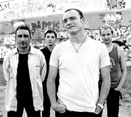 A photo of members of Louise Attaque standing outside in front of a wall covered with graffiti.